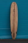 The back of this wooden surfboard also displays the bizia brand name and bird logo near the nose. The three-fin set up is also visible with a large longboard fin slot bordered by two smaller fin slots on either side.