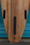 A close-up of the back of the wooden board's squash tail shows a large longboard fin slot with two smaller fin slots on either side. At the bottom is an albizia project certification logo, signifying the use of albizia wood as regenerative lumber to make this sustainable surfboard.