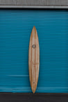 A large and imposing wooden surf board boasts the legendary Dick Brewer logo. This gun surfboard is long and narrow with both a pointed tail and nose.