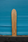 Wooden alaia surfboard with an iwa bird, the signature of shaper Chris Miyashiro, engraved near the bottom. The board is long and thin, curving softly towards the flat tail.
